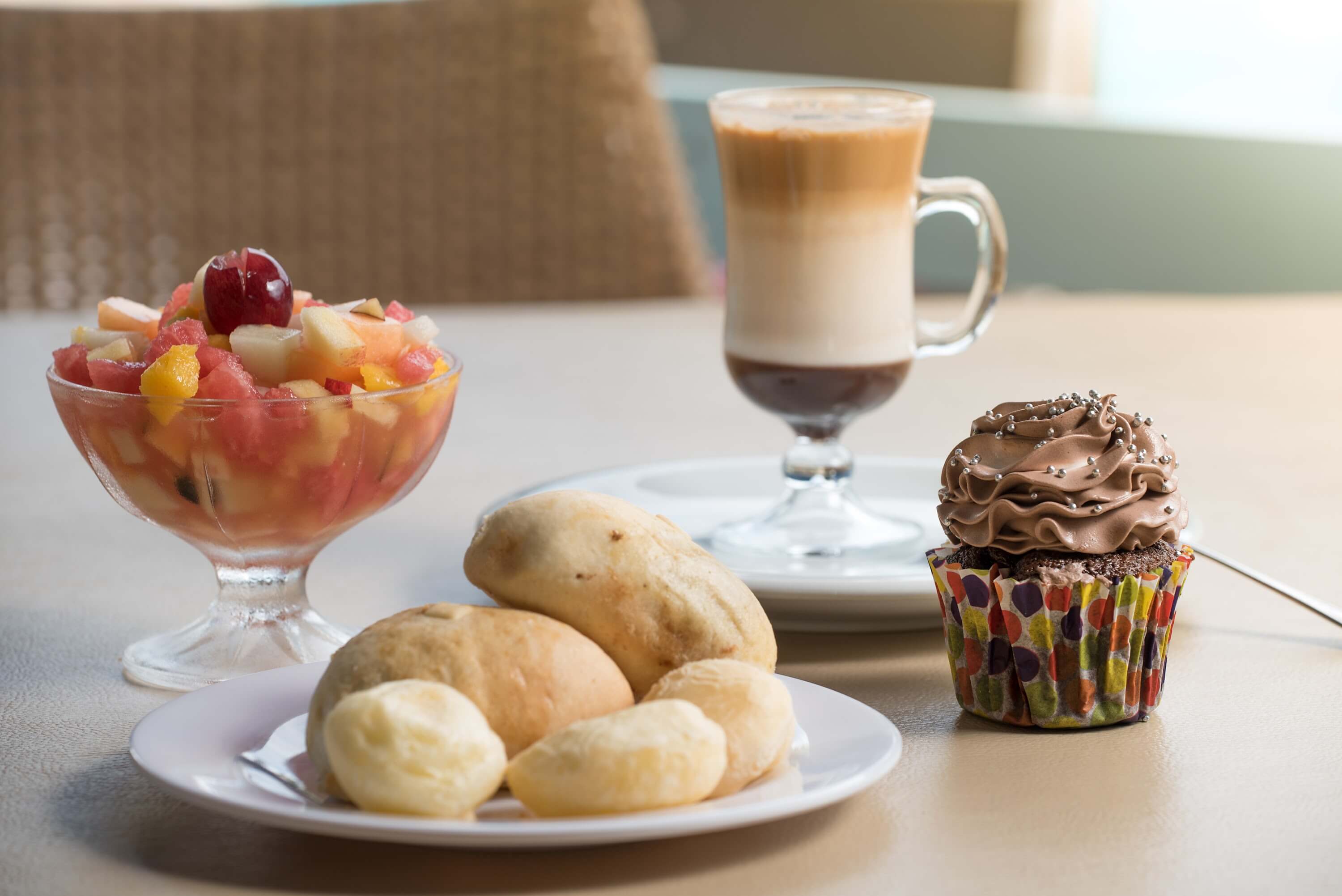 Salada de furtas, prato com pães de queijo, cupcake de chocolate decorado e uma xícara com café mocha ao fundo, todos os itens sobre a mesa, no Espaço Canoas.